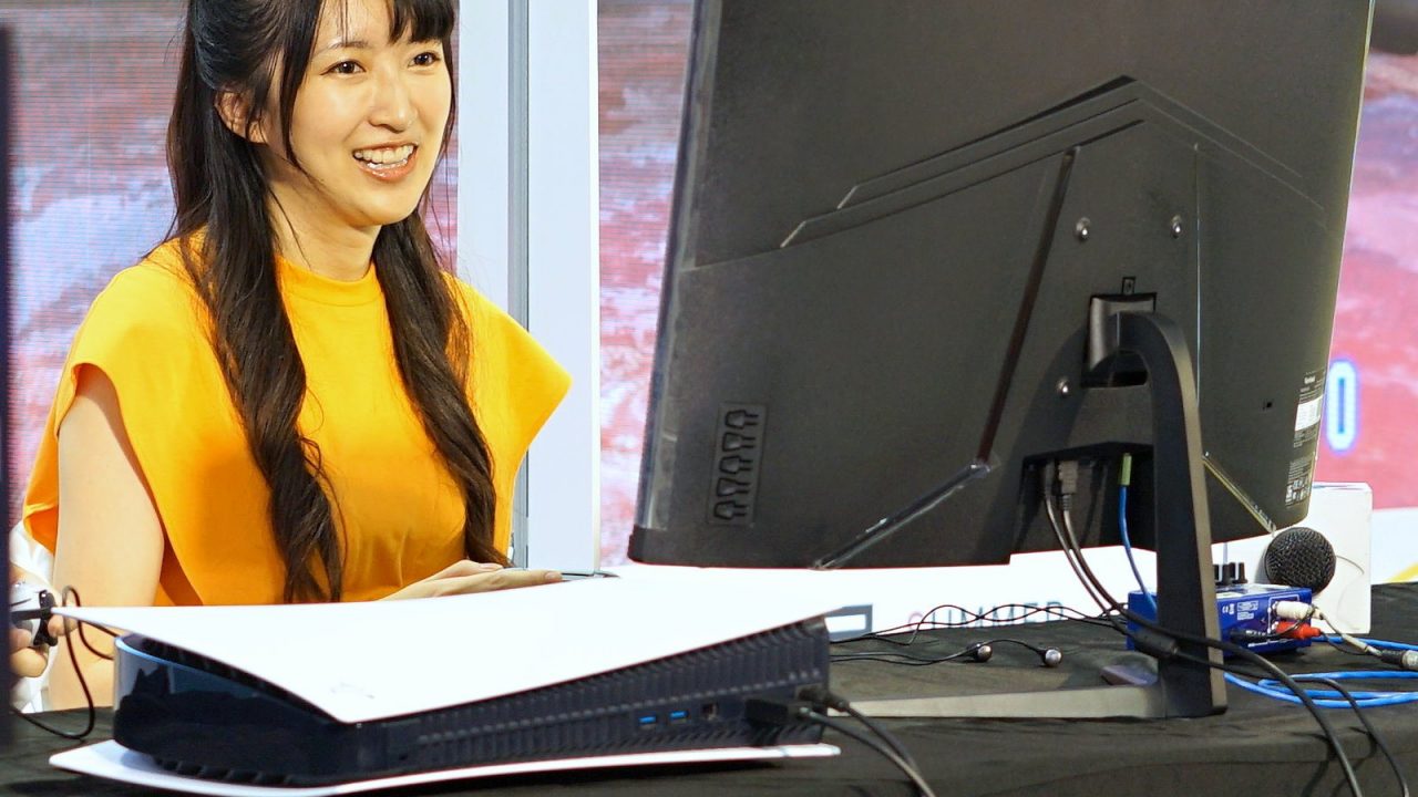 PS5 lying horizontally beside a monitor on a stage, with a presenter smiling